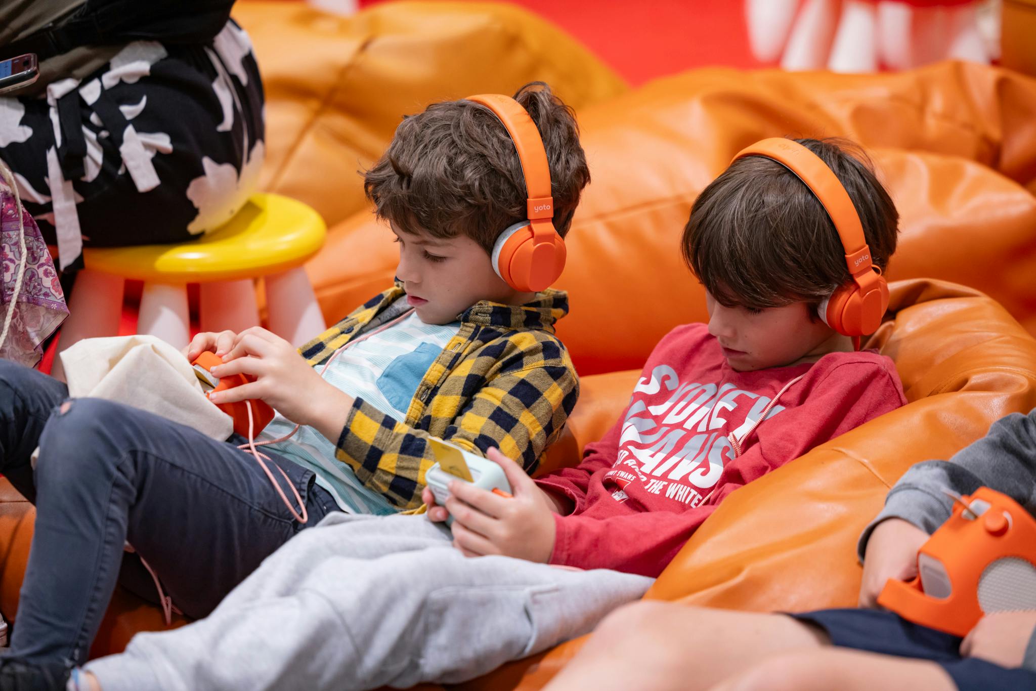 Two kids, sitting on a comfy orange beanbag, wearing Yoto headphones and listening to Yoto Minis.