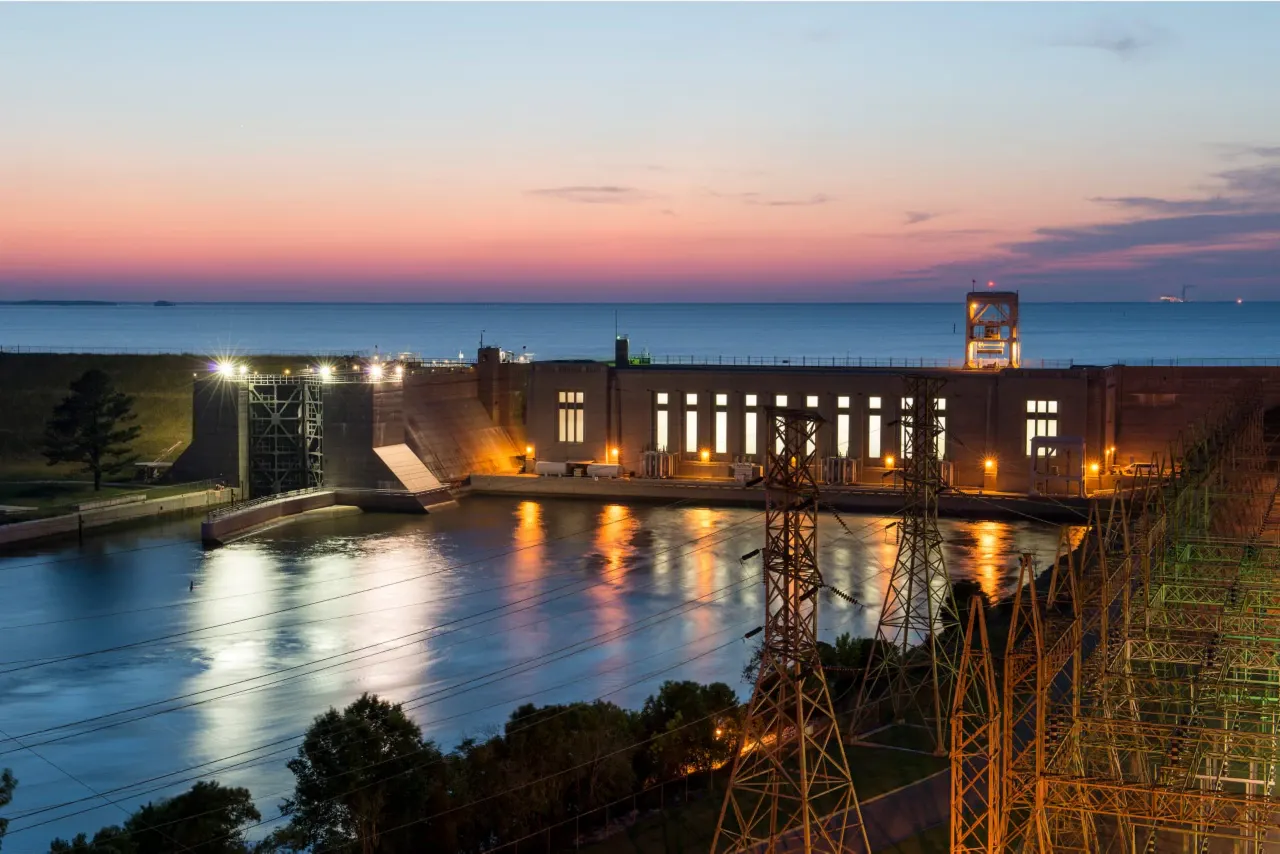 A large concrete dam at sunset. The dam is located on a river and there is a body of water behind it. Power lines are running along the side of the dam.
