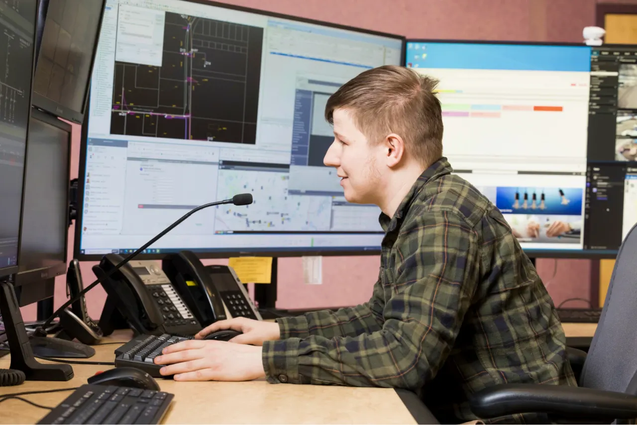 A man in a plaid shirt sits at a desk, monitoring multiple computer screens displaying maps and data