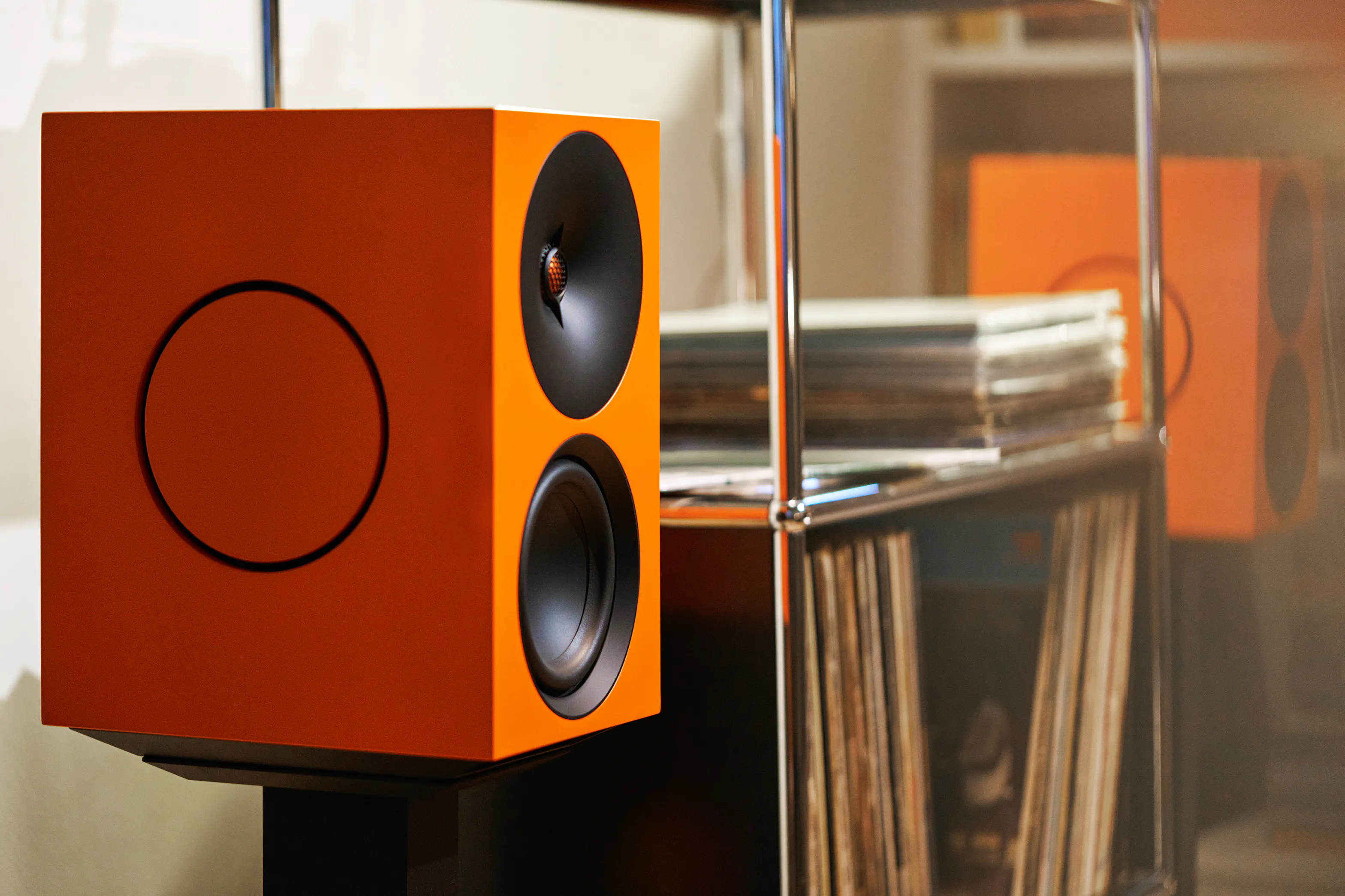 Close up of an L/R X active speaker in orange, with a second speaker and vinyl storage behind.