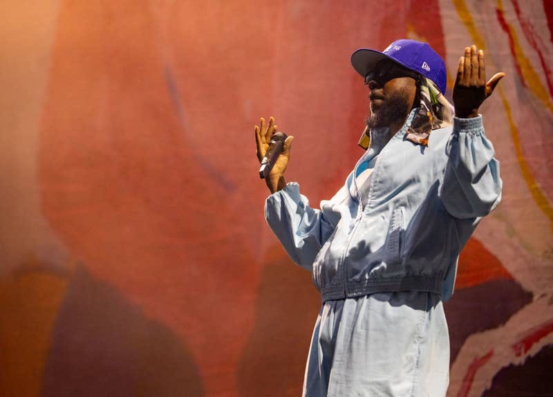 Music artist performing on stage with arms raised, wearing casual attire with a hat