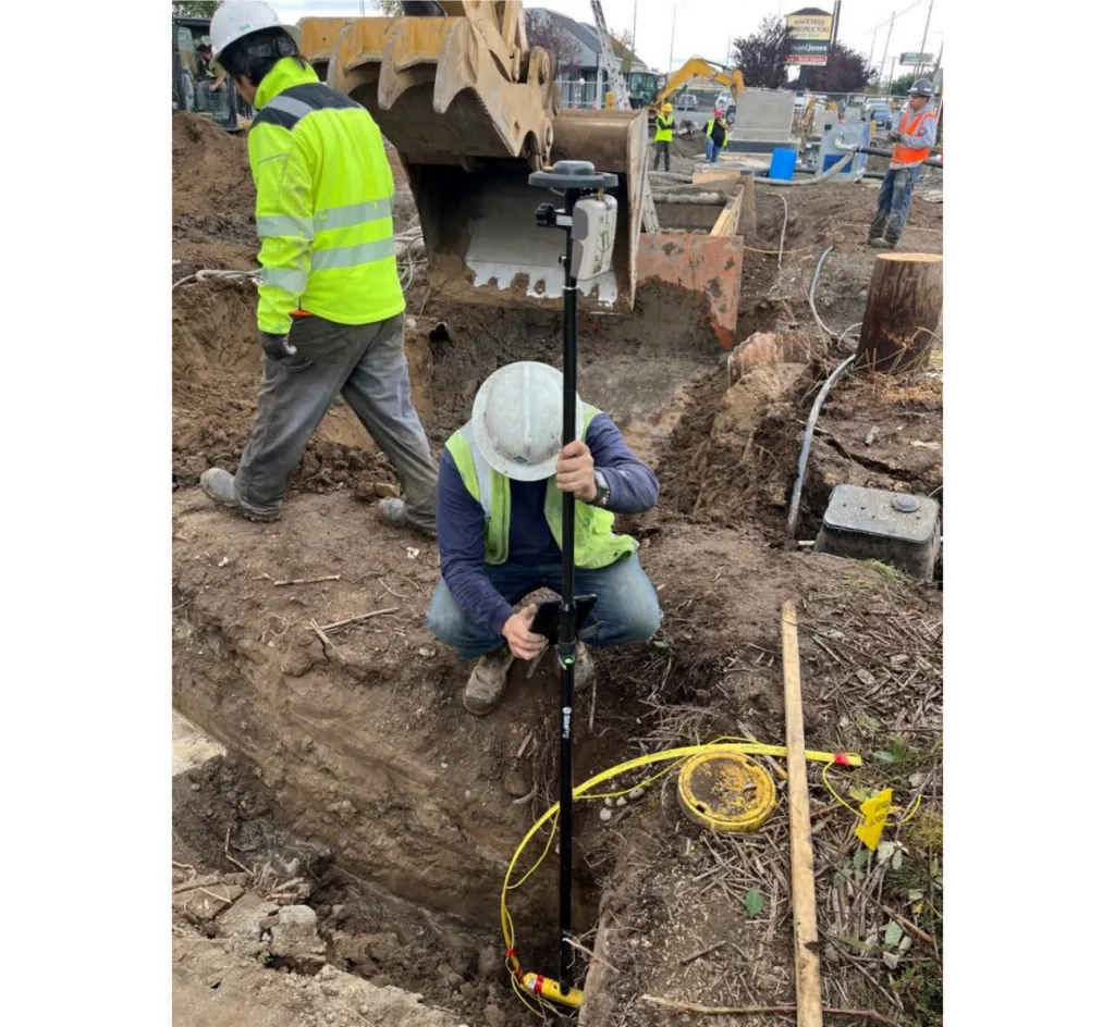Construction workers surveying at a trench site; One uses a pole-mounted device near a yellow utility cover